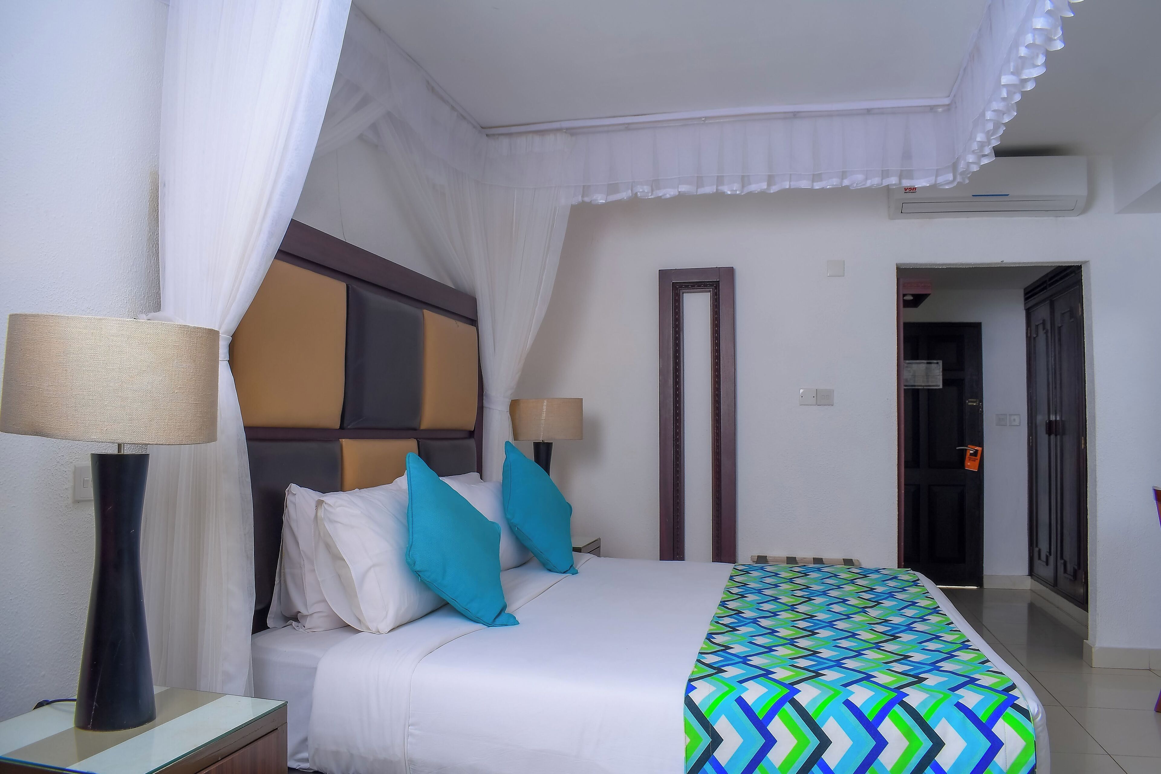 A hotel room with a bed, two bedside lamps, a patterned bedspread, and a door leading to another room.