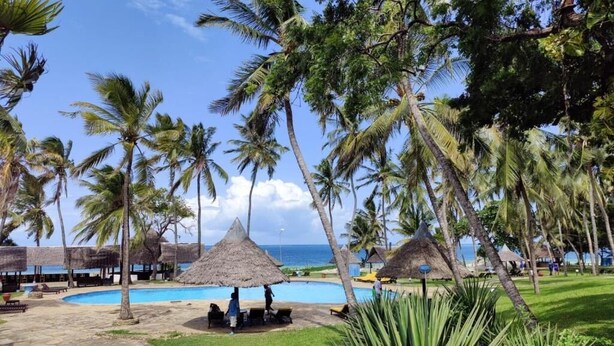 A pool area with palm trees, a thatched-roof structure, and a clear view of the ocean. - Cocoa Luxury Resort, Nyali (Mombasa)