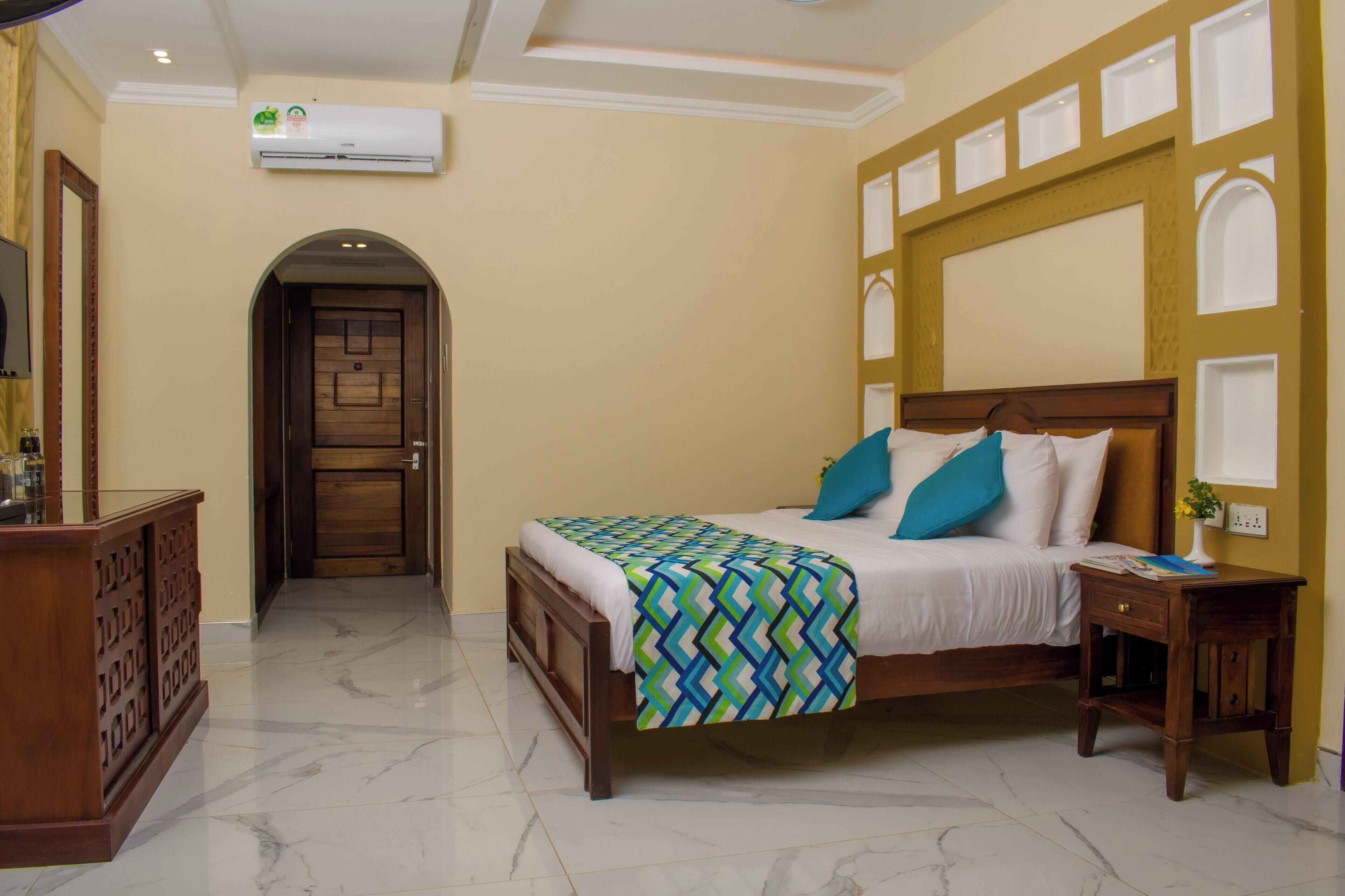 A hotel room with a bed, a nightstand, a desk, an air conditioning unit, and a door leading to another room.