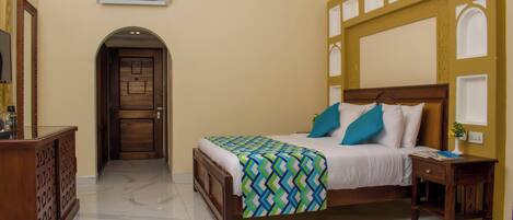A hotel room with a bed, a nightstand, a desk, an air conditioning unit, and a door leading to another room.