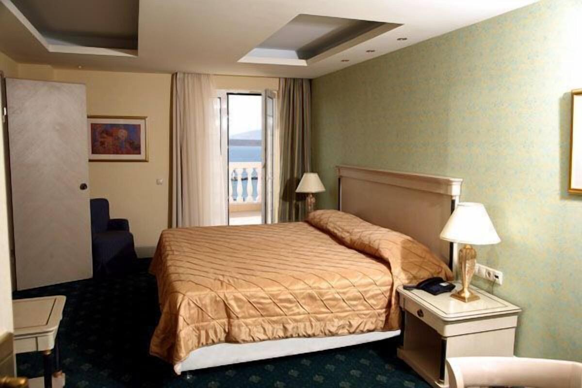 A hotel room with a bed, a desk, a chair, a nightstand with a lamp, and a view of the sea through a window.
