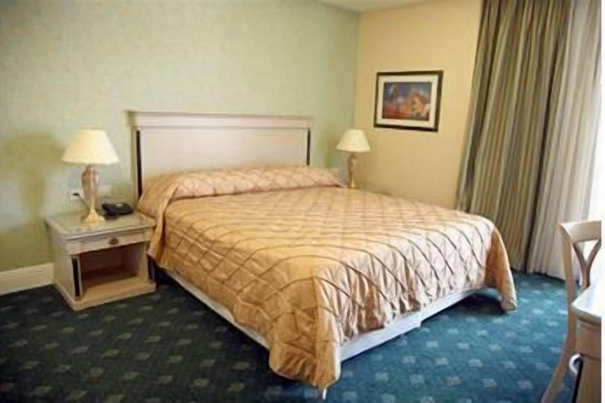 A hotel room with a bed, two bedside tables with lamps, a chair, a framed picture on the wall, and a window with curtains.