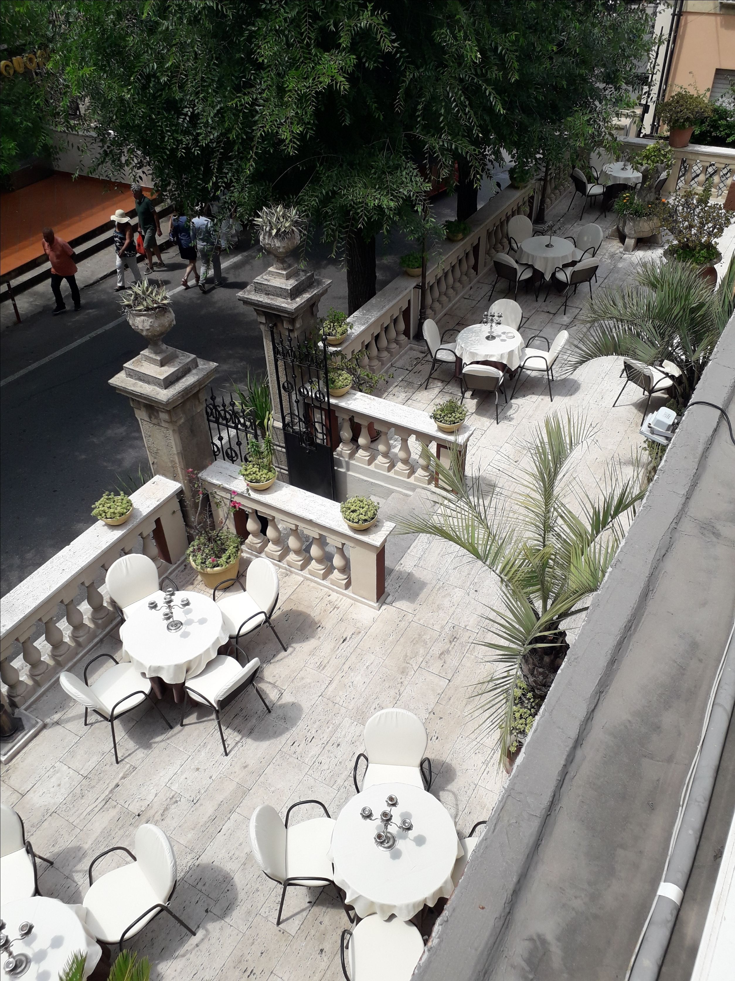 Un café al aire libre con mesas redondas y sillas blancas, rodeado de macetas con plantas y un balcón de piedra.