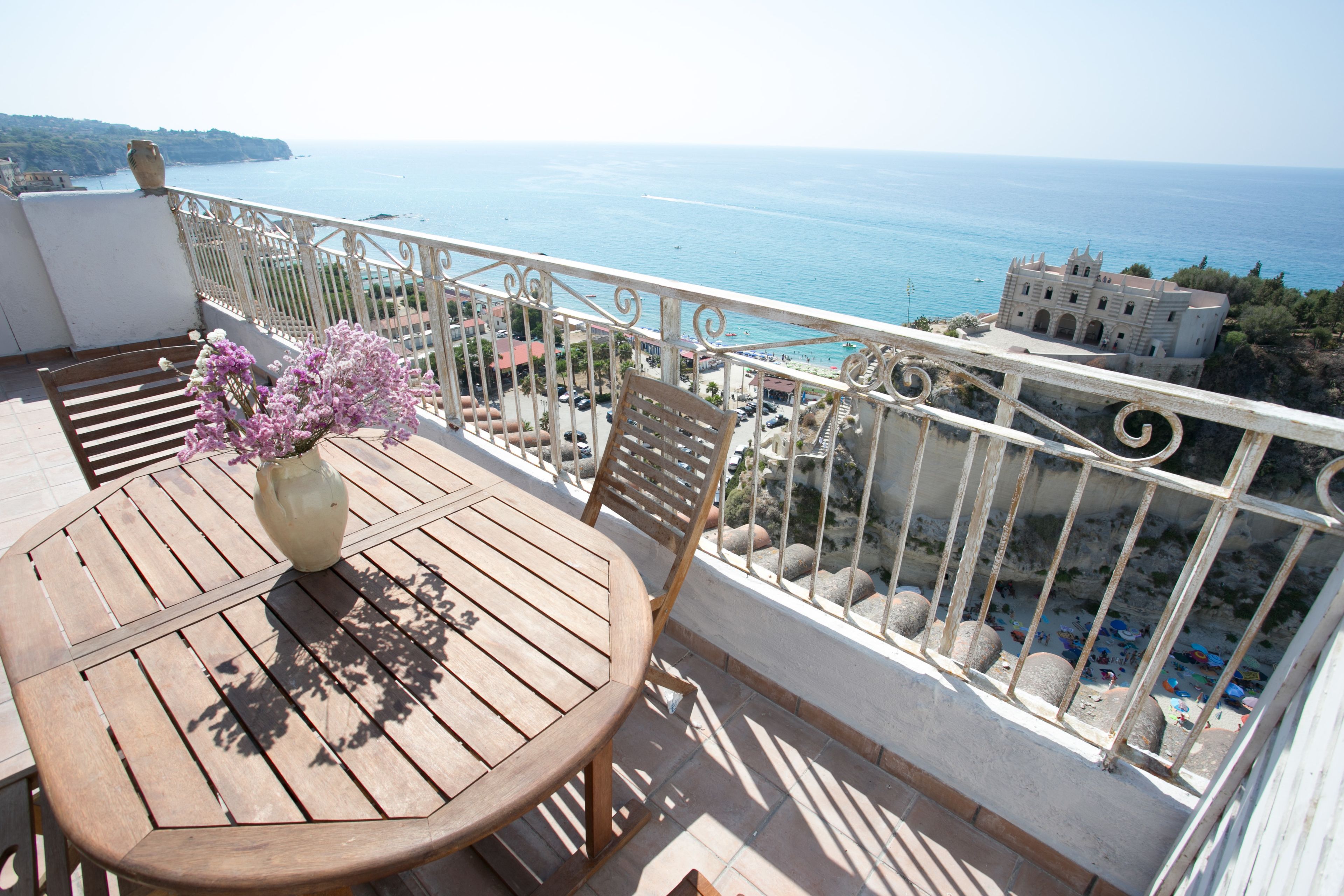 A wooden table with a vase of flowers on a balcony overlooking a coastal view.