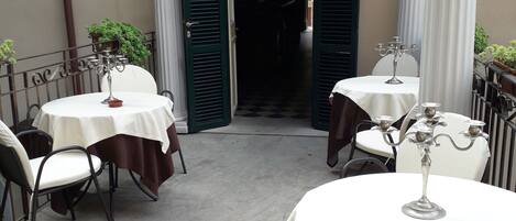 Una terrazza esterna con tavoli e sedie rotonde, una porta con persiane verdi e una pianta in vaso.