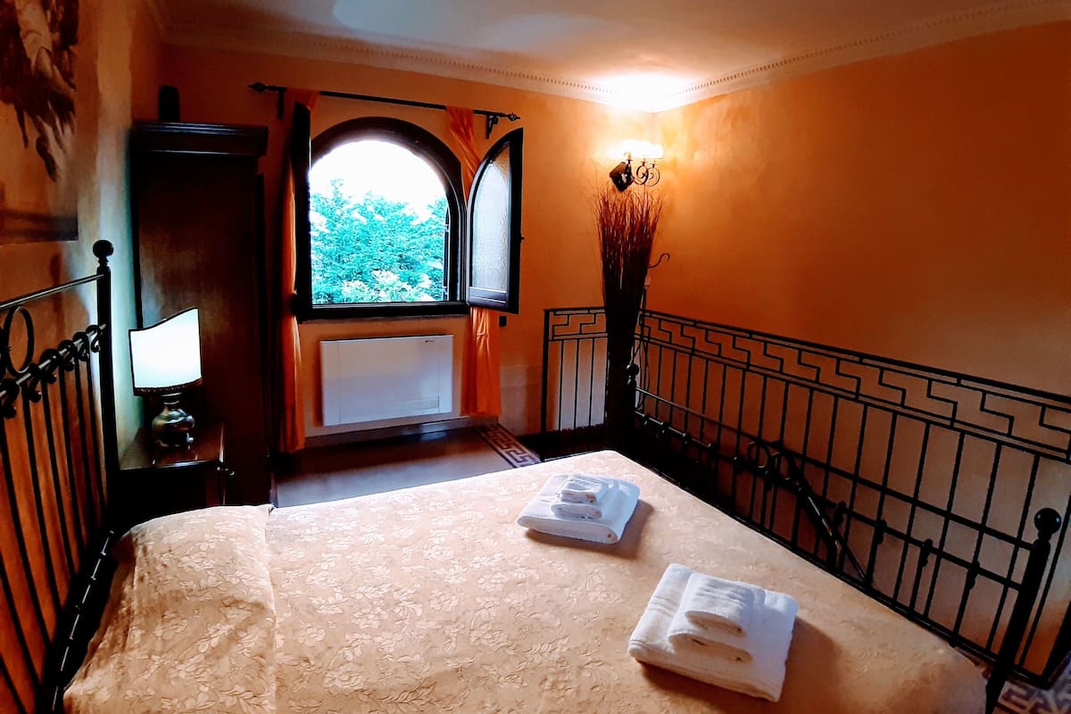 A hotel room with a bed, a radiator, a window with curtains, a lamp, and a vase with branches.