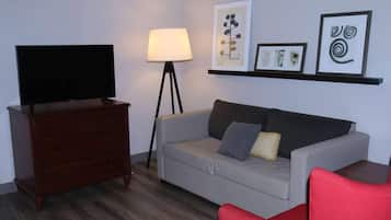 A living room with a television on a wooden dresser, a grey sofa with cushions, a floor lamp, and framed artwork on the wall.