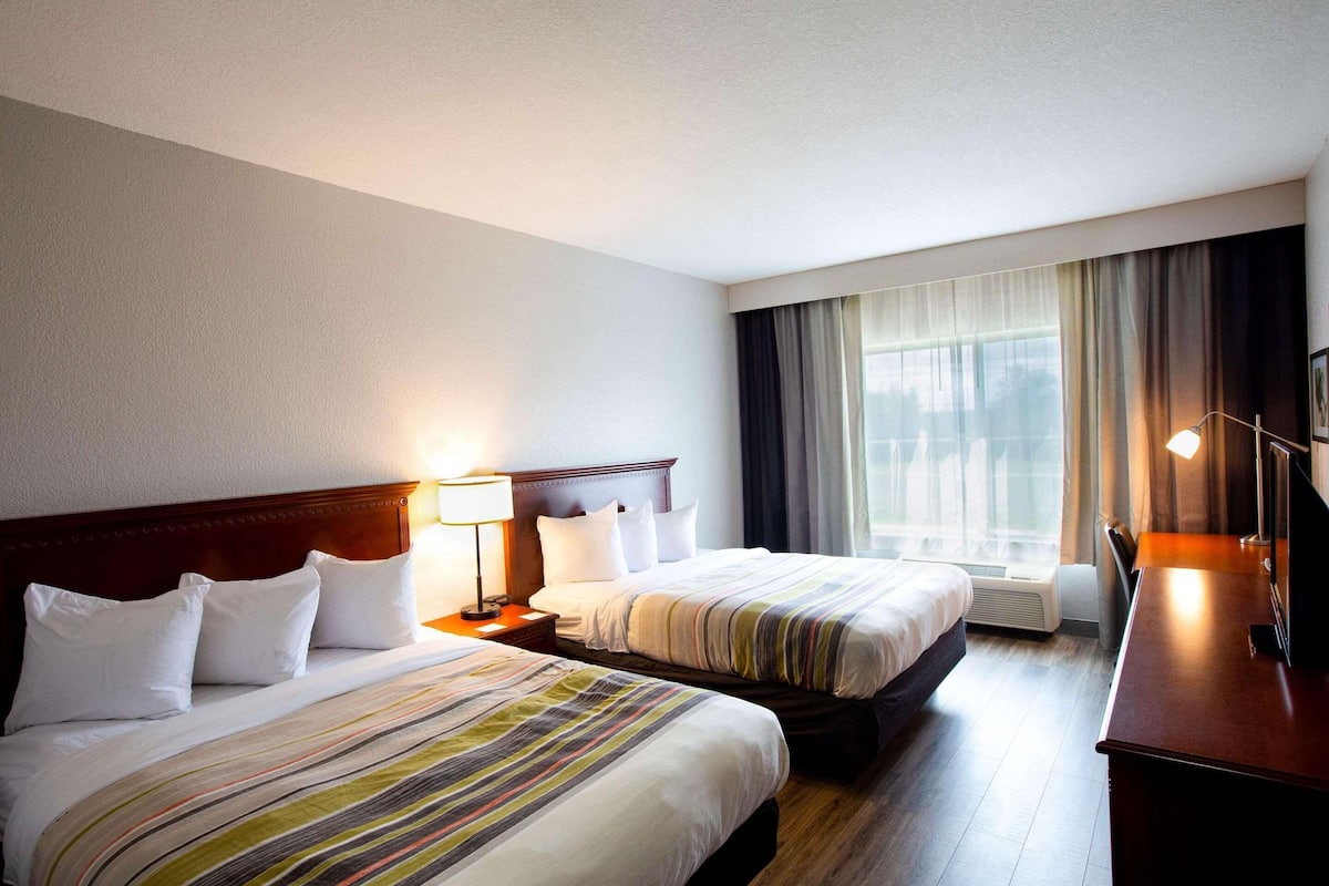 A hotel room with two beds, a wooden headboard, a desk with a lamp, and a large window with curtains.