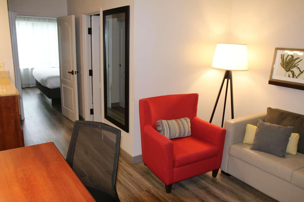 A hotel room with a wooden dining table, a red armchair, a white sofa, a floor lamp, and a framed picture on the wall.