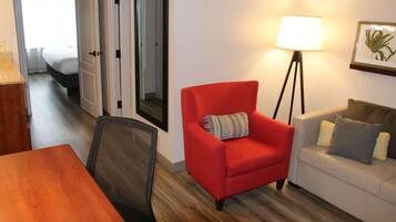 A hotel room with a wooden dining table, a red armchair, a white sofa, a floor lamp, and a framed picture on the wall.