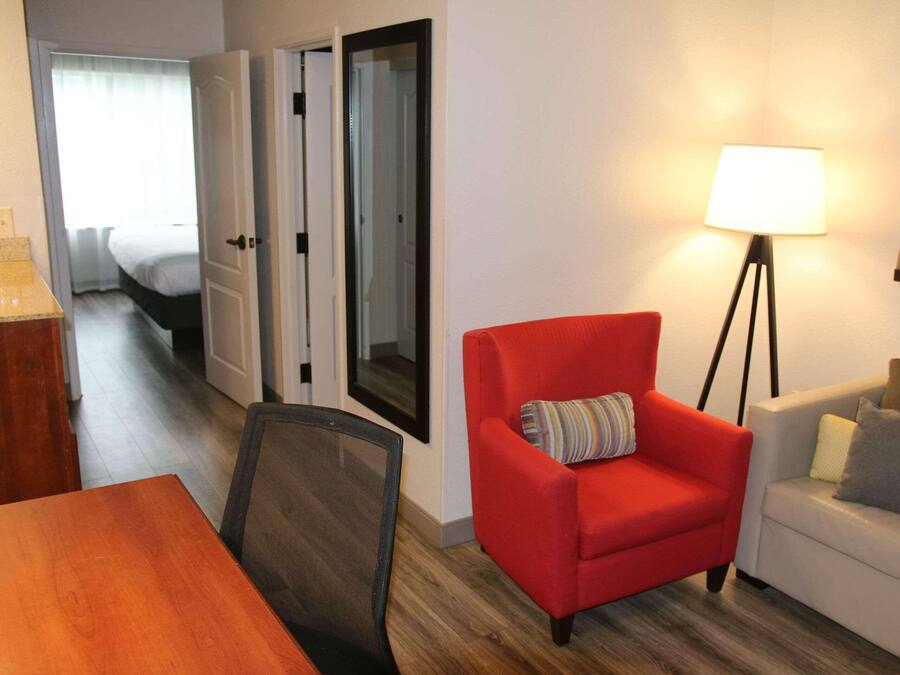 A hotel room with a wooden dining table, a red armchair, a white sofa, a floor lamp, and a framed picture on the wall.