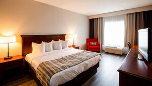 A hotel room with a large bed, two bedside tables with lamps, a red armchair, a desk with a television, and a window with curtains. - Country Inn & Suites by Radisson, Wilson, NC (Wilson)
