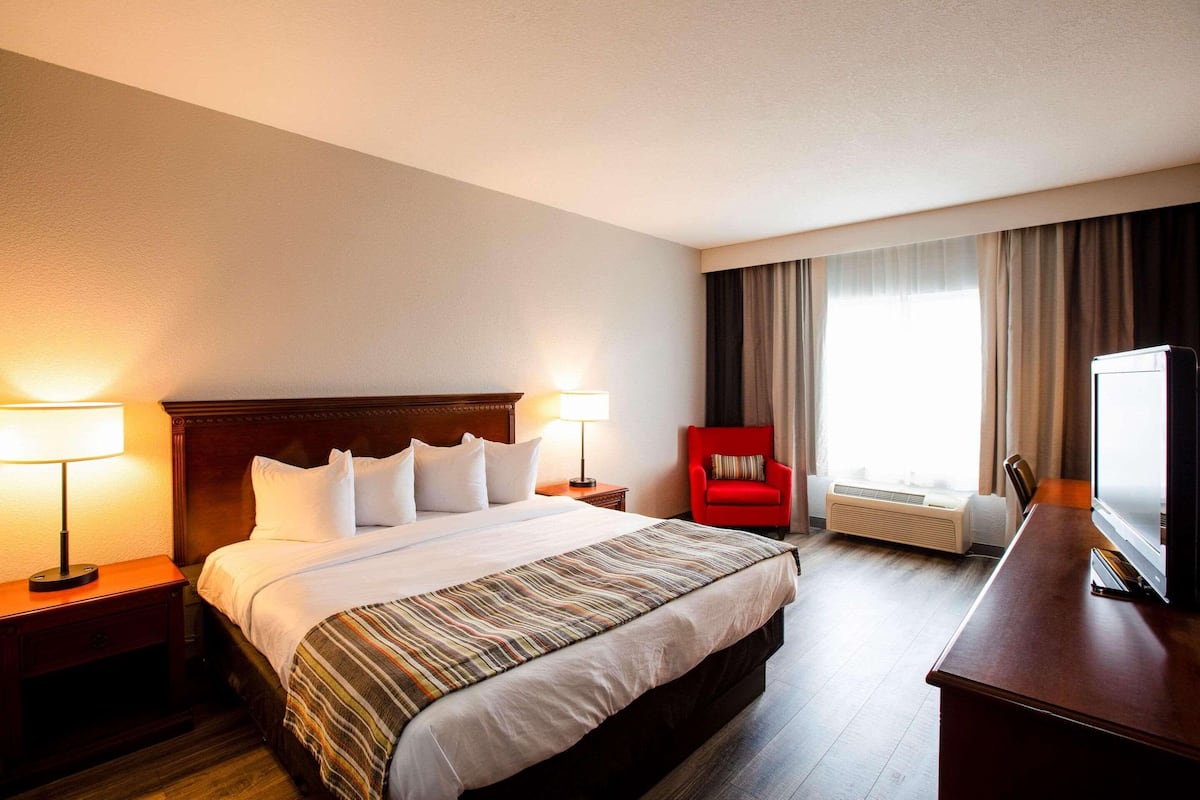 A hotel room with a large bed, two bedside tables with lamps, a red armchair, a desk with a television, and a window with curtains.
