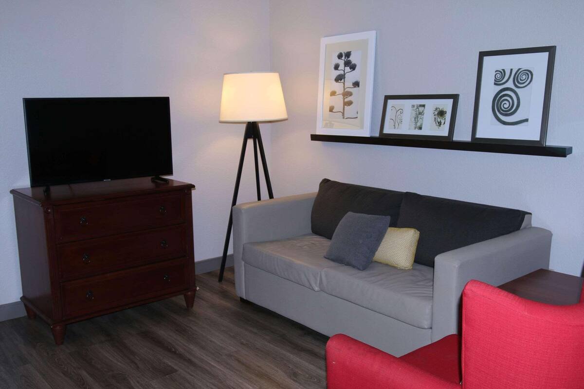 A living room with a flat-screen TV, a wooden dresser, a grey sofa with cushions, a red armchair, a floor lamp, and framed artwork on the wall.