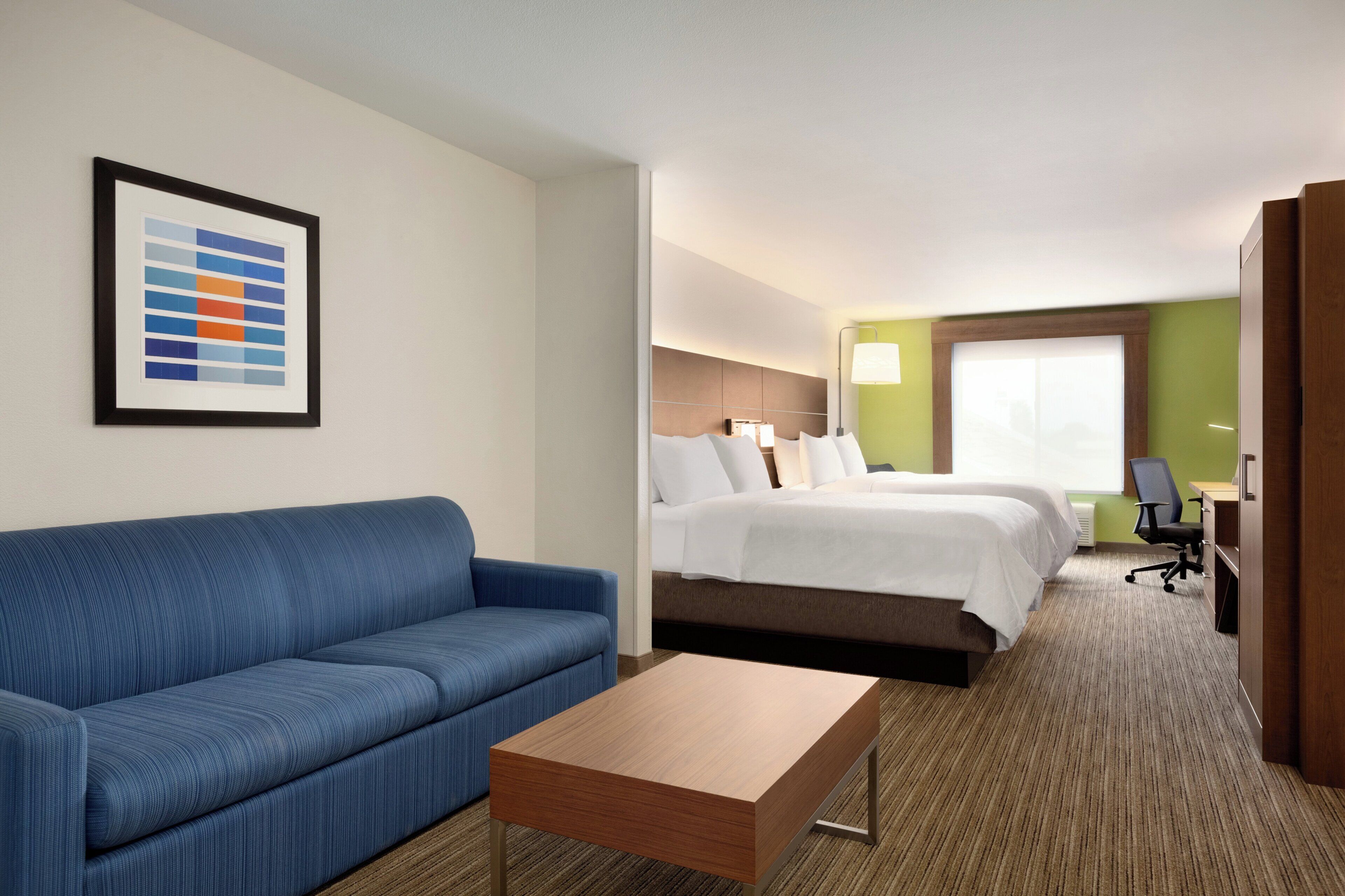 Habitación de hotel con sofá azul, una mesa de centro de madera, cama con ropa blanca, escritorio con silla y una tabla de colores enmarcada en la pared.