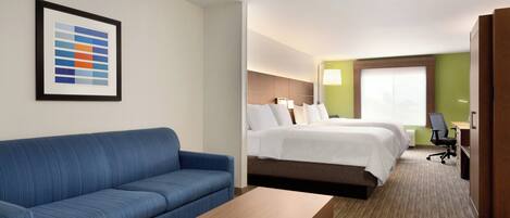 Habitación de hotel con sofá azul, una mesa de centro de madera, cama con ropa blanca, escritorio con silla y una tabla de colores enmarcada en la pared.