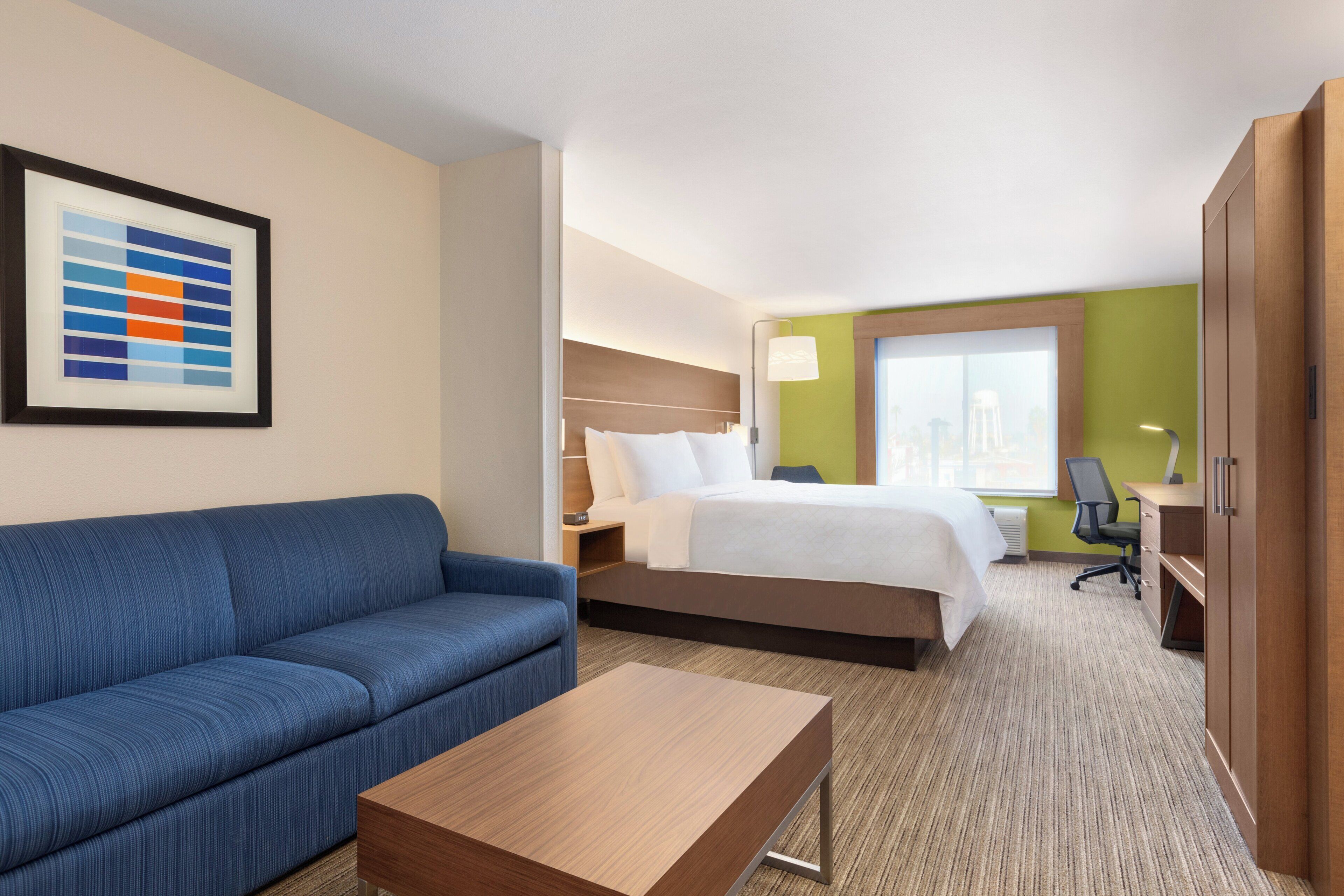 A hotel room with a blue sofa, a wooden coffee table, a bed with white bedding, a desk with a chair, and a large window.