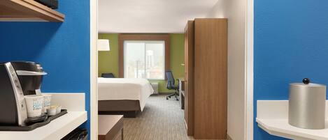 Habitación de hotel con cama, escritorio con cafetera, lámpara y vista a la ciudad a través del ventanal.