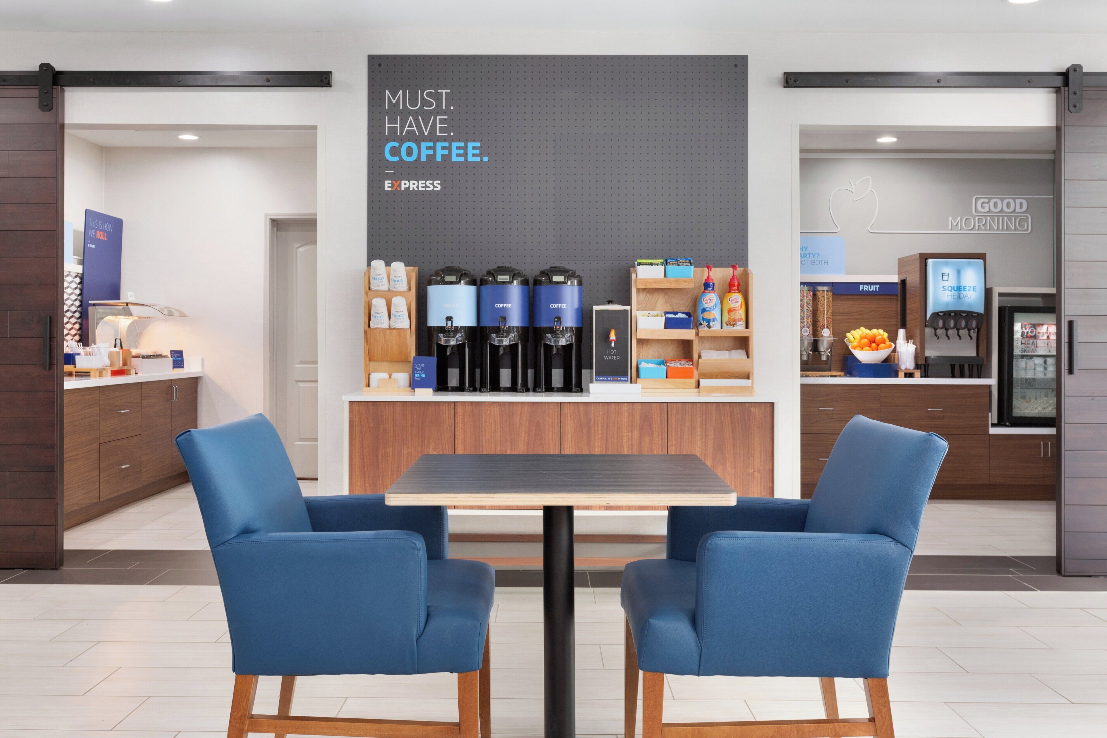 Een moderne koffiehoek met koffieautomaten met branding, een zithoek met twee blauwe stoelen en een muur met inspirerende tekst.