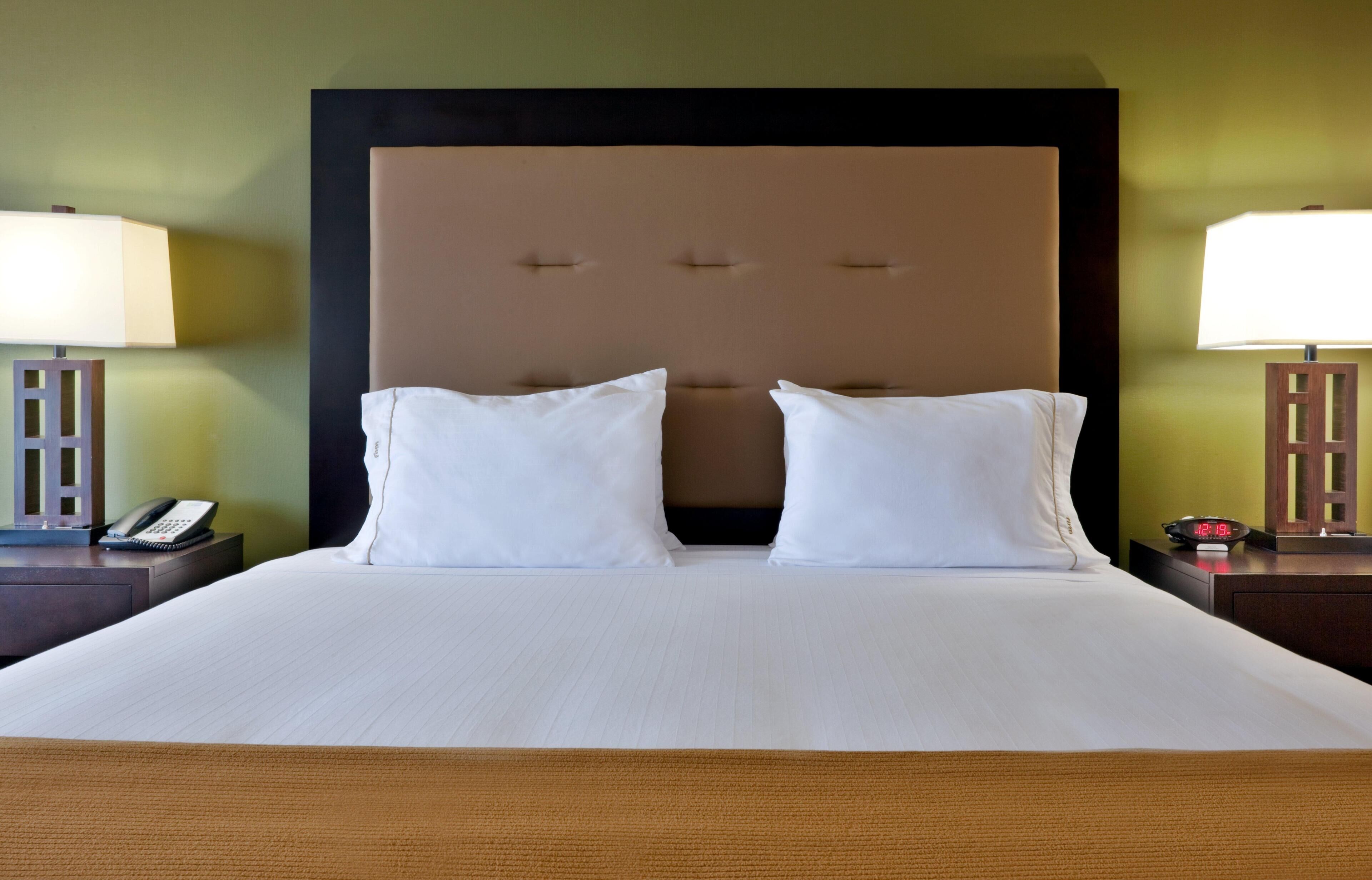 A hotel room with a large bed, two bedside tables, a nightstand, a lamp, a phone, and a digital clock.
