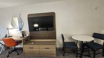 A hotel room with a wooden desk, a chair, a small table with two chairs, a mirror, and a lamp.