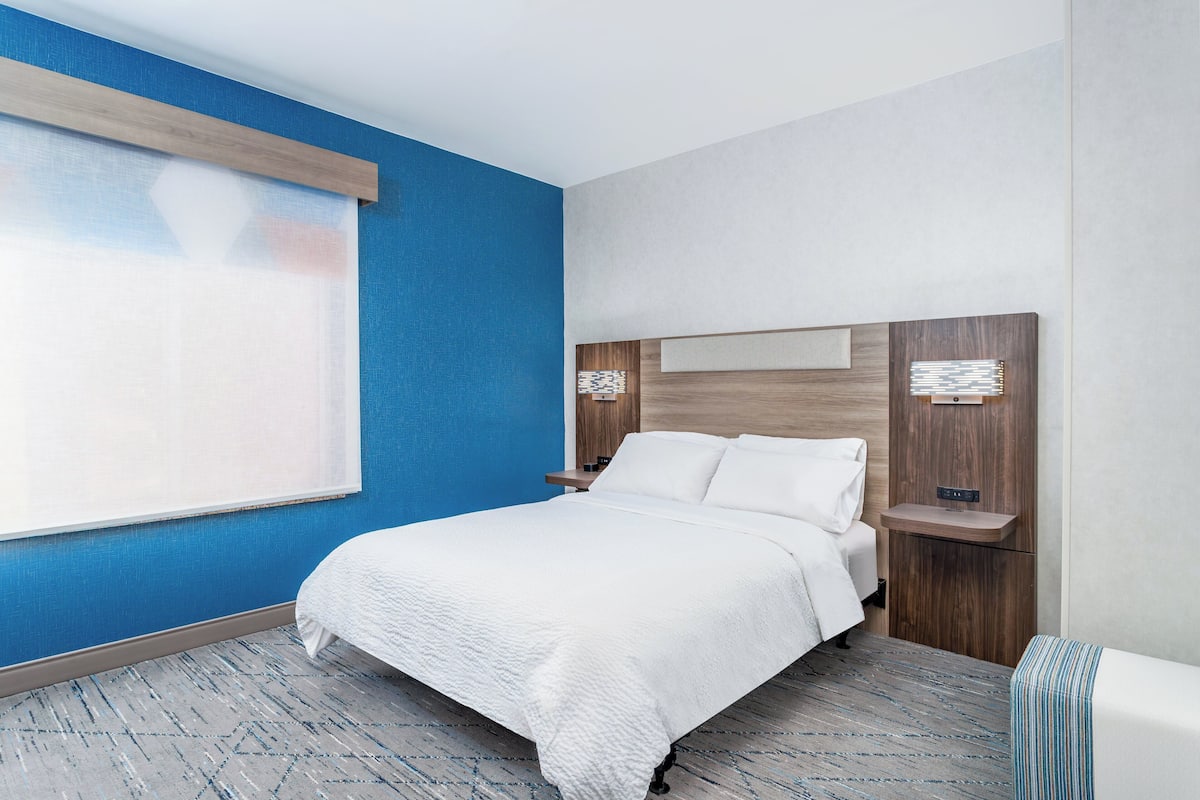 A modern hotel room with a large bed, a nightstand, a wall-mounted light, and a window with blinds.