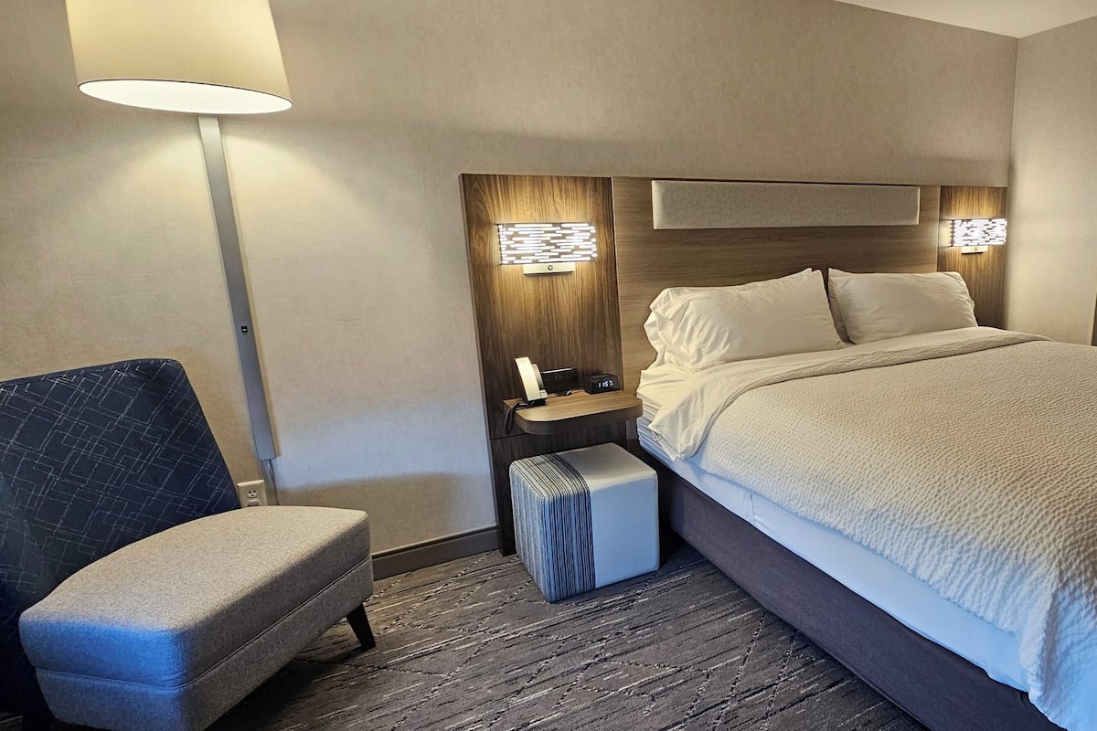 A hotel room with a large bed, a bedside table with a lamp, a chair, and a small ottoman.