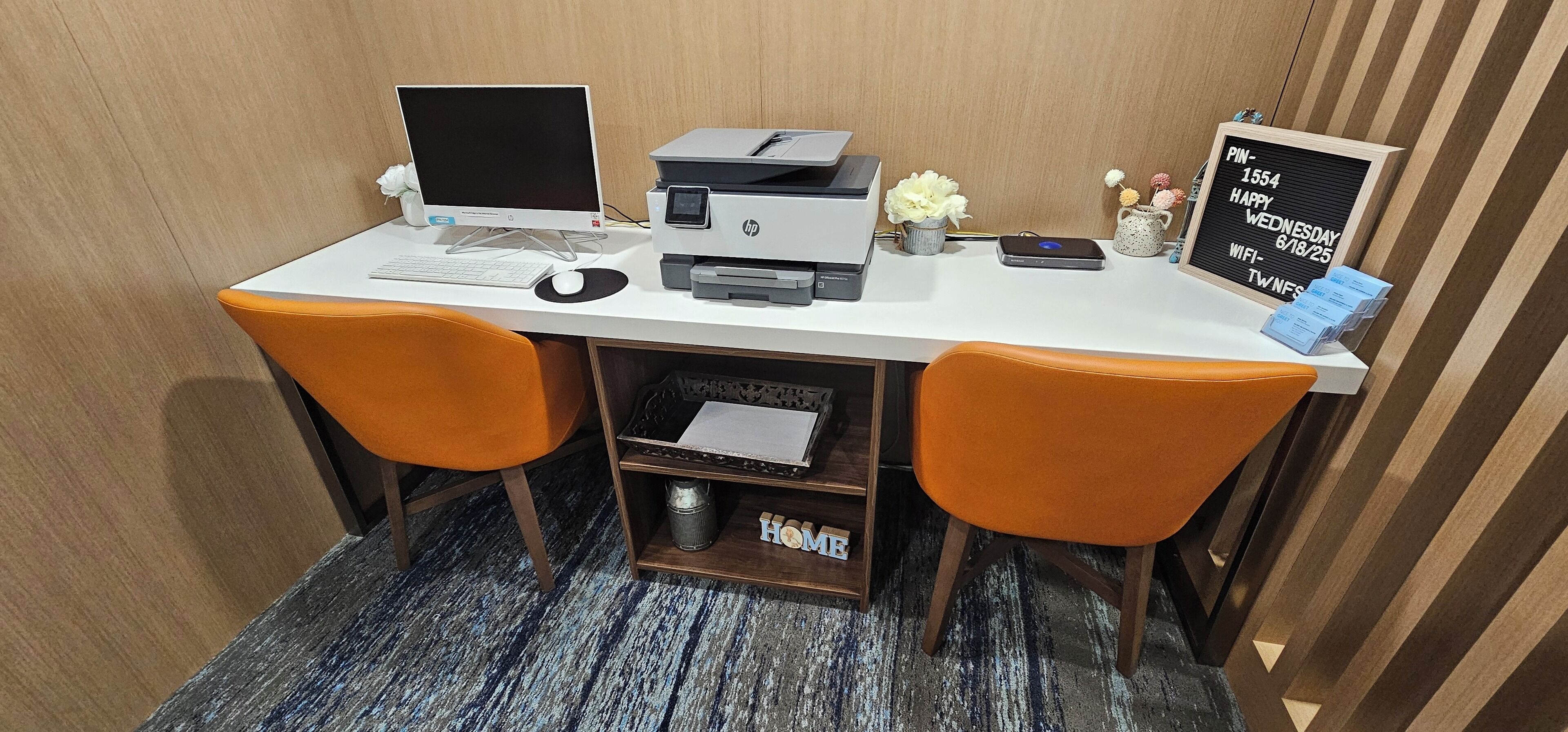 A hotel workspace with two orange chairs, a desk with a computer, printer, and a signboard.