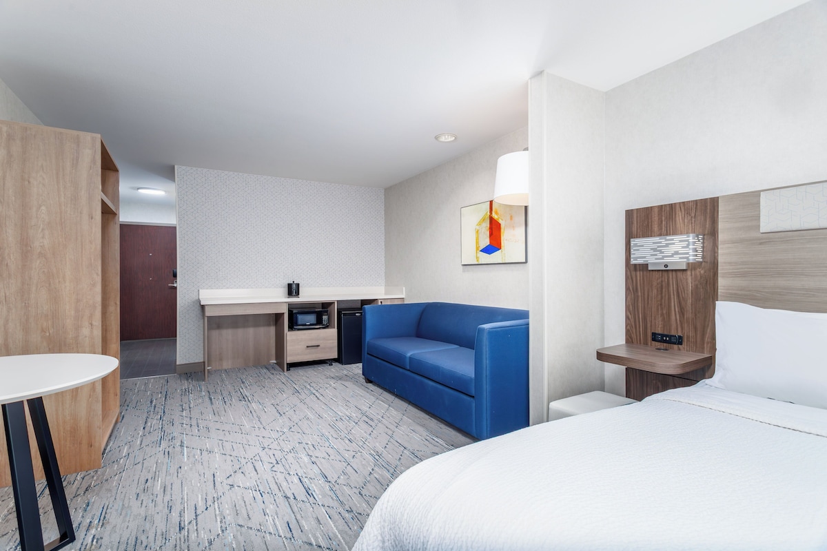 Habitación de hotel con una cama, un sofá azul, un escritorio con microondas, una mesita pequeña y un televisor montado en la pared.