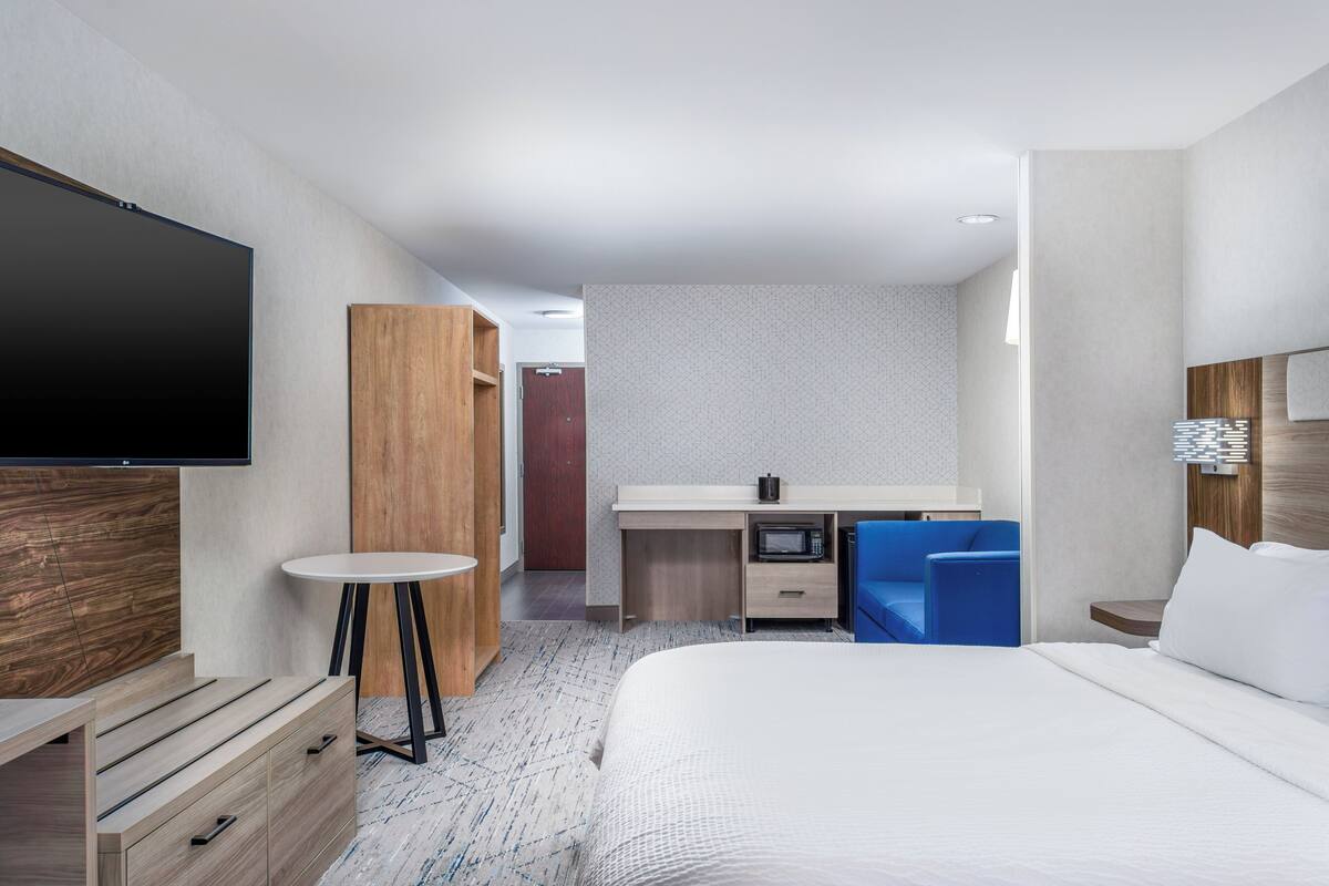 A hotel room with a bed, a TV, a desk with a microwave, a chair, and a wardrobe.