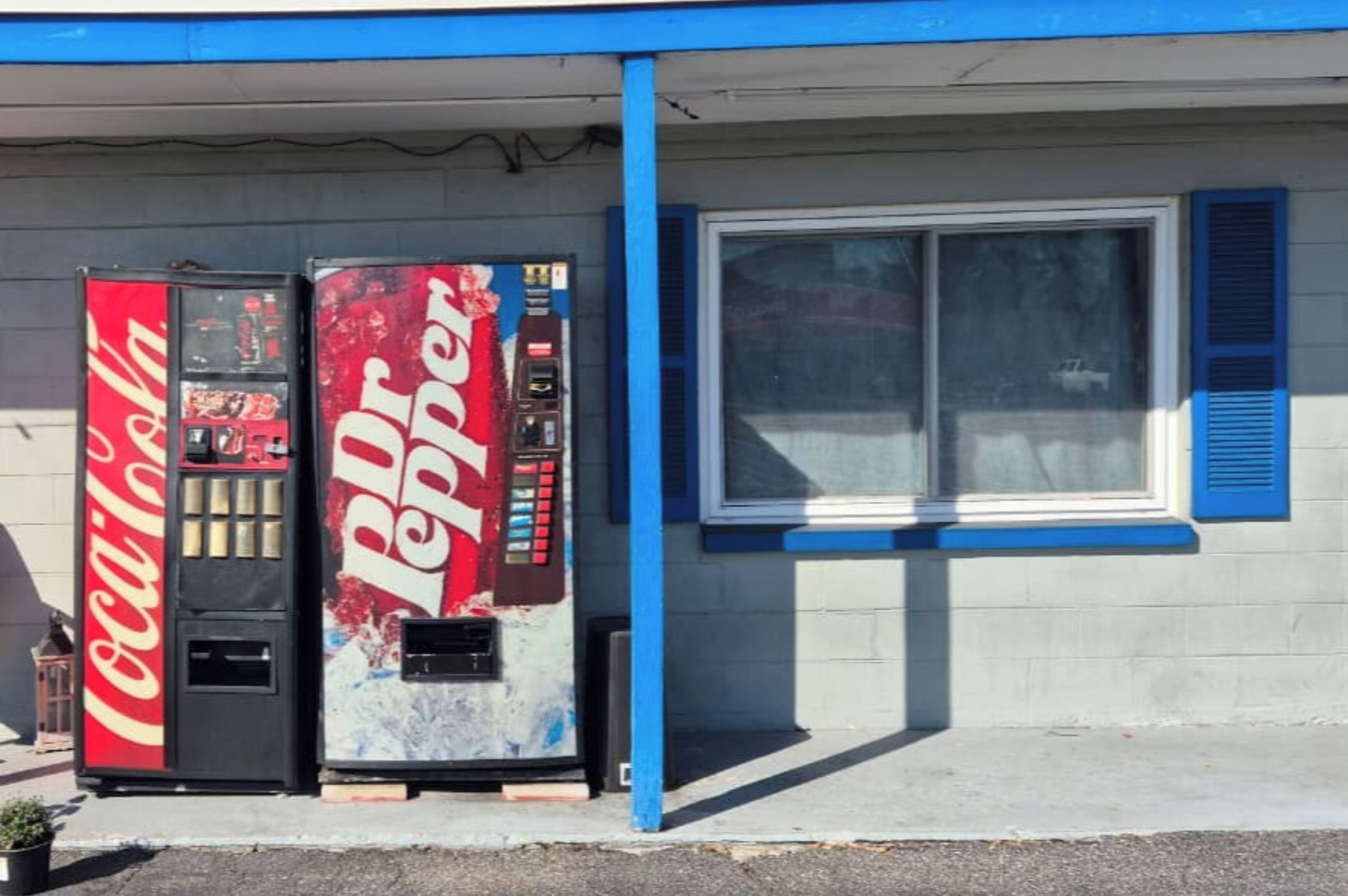 Two vending machines, one Coca-Cola and one Dr Pepper, in front of a building with a blue trim and a window.