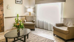 A hotel room with two beige armchairs, a round coffee table, a lamp, a large window with curtains, and a framed picture on the wall. - Sleep Inn & Suites Gonzales near Palmetto State Park (Gonzales)