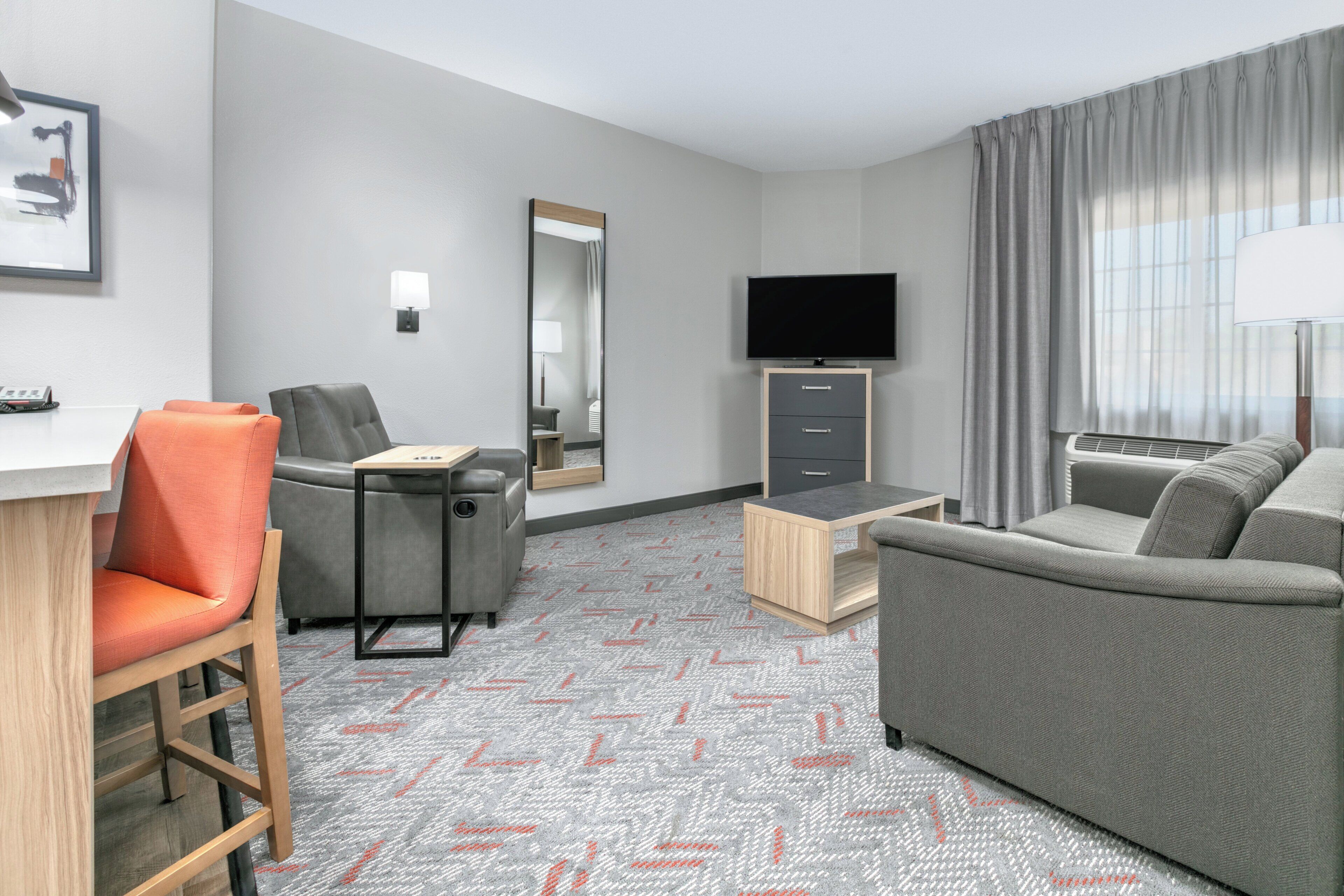 A hotel room with a desk, two chairs, a sofa, a television, a mirror, and a window with curtains.