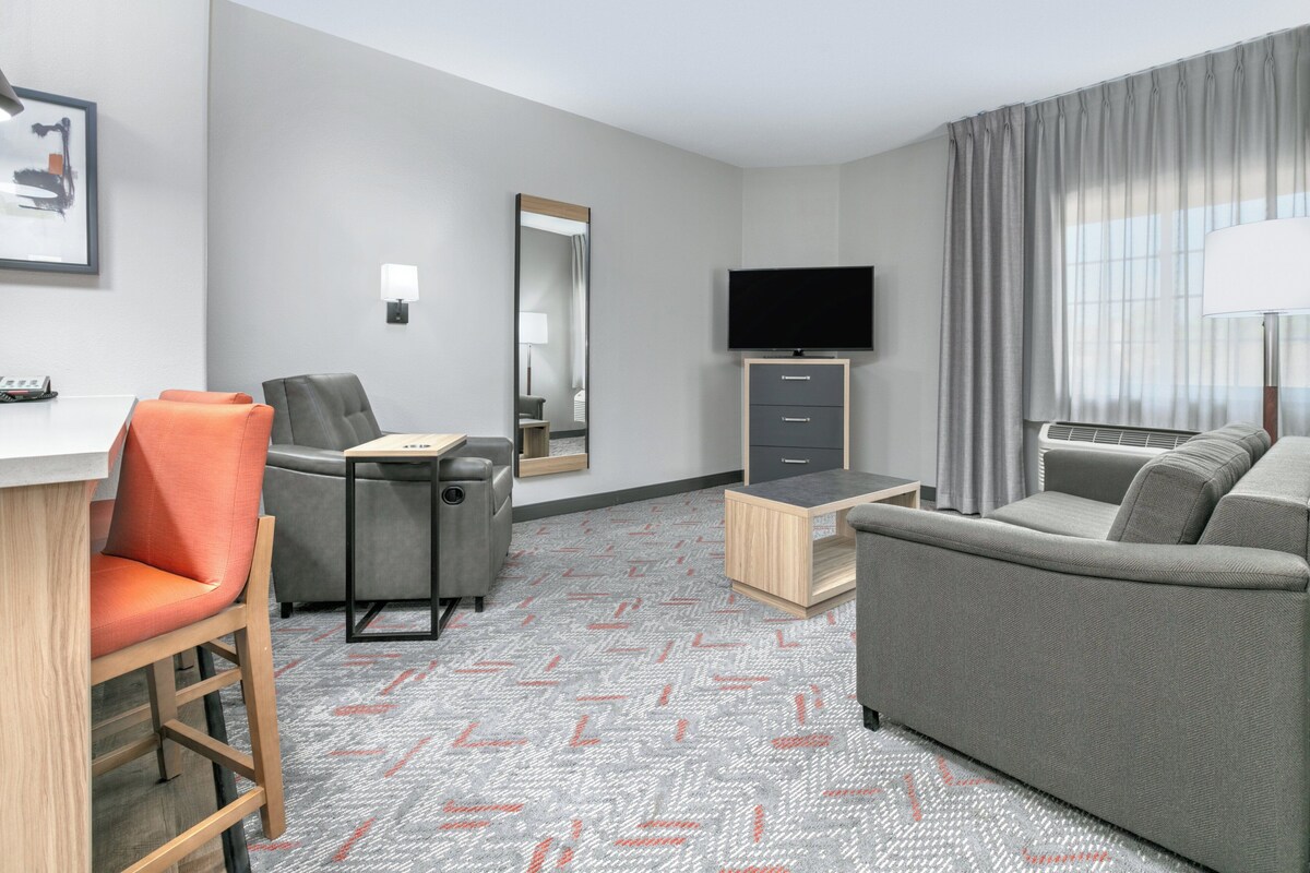 Habitación de hotel con un sofá gris, un sillón gris, una mesita de madera, un televisor y un espejo.