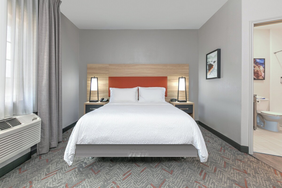 Habitación de hotel con una cama grande, cabecera de madera, dos mesitas de noche con lámparas, un radiador y un baño visible a través de una puerta abierta.