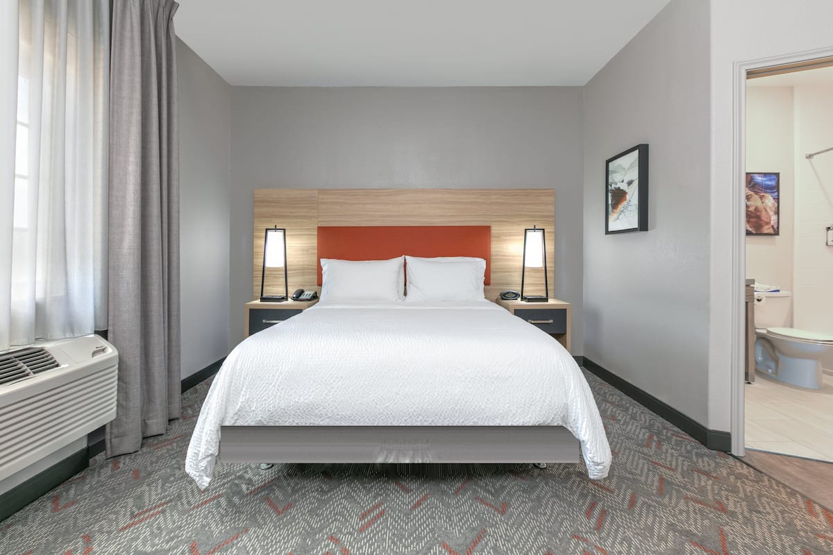 Habitación de hotel con una cama grande, cabecera de madera, dos mesitas de noche con lámparas, un radiador y un baño visible a través de una puerta abierta.