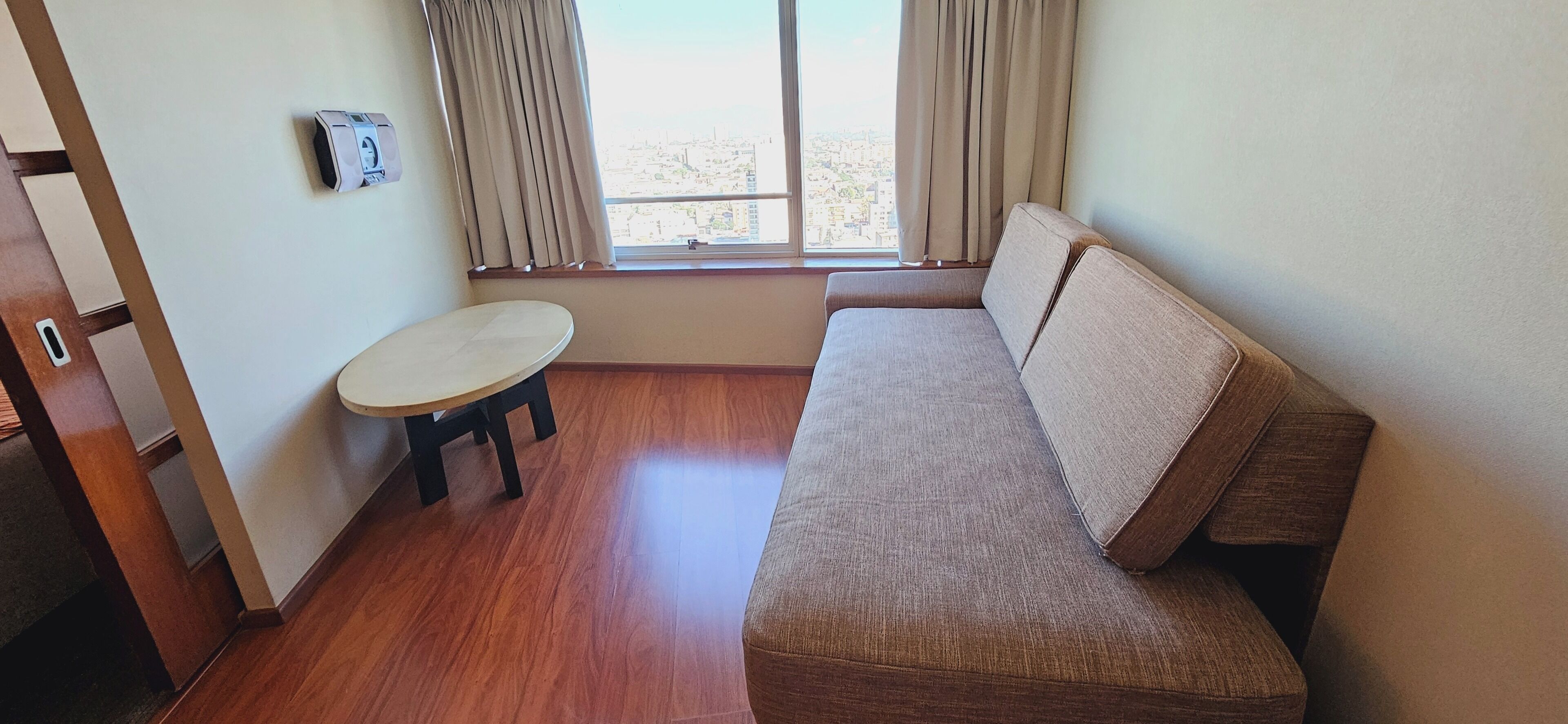 Una habitación con sofá, una mesita redonda, ventana con cortinas y aire acondicionado de pared.