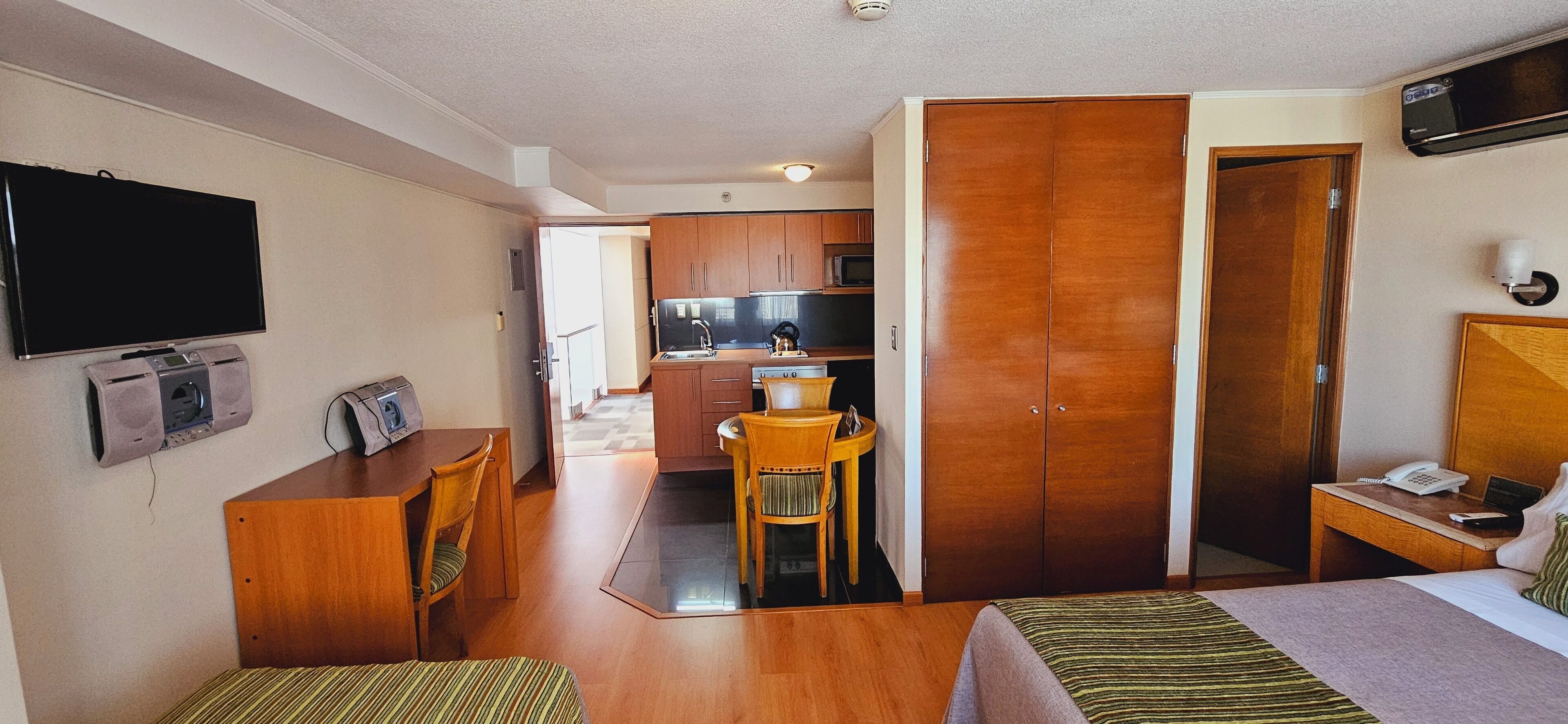 Habitación de hotel con cama, escritorio, teléfono, televisor y una kitchenette al fondo.