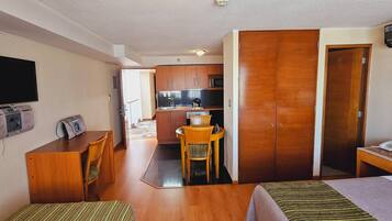 Habitación de hotel con cama, escritorio, teléfono, televisor y una kitchenette al fondo.