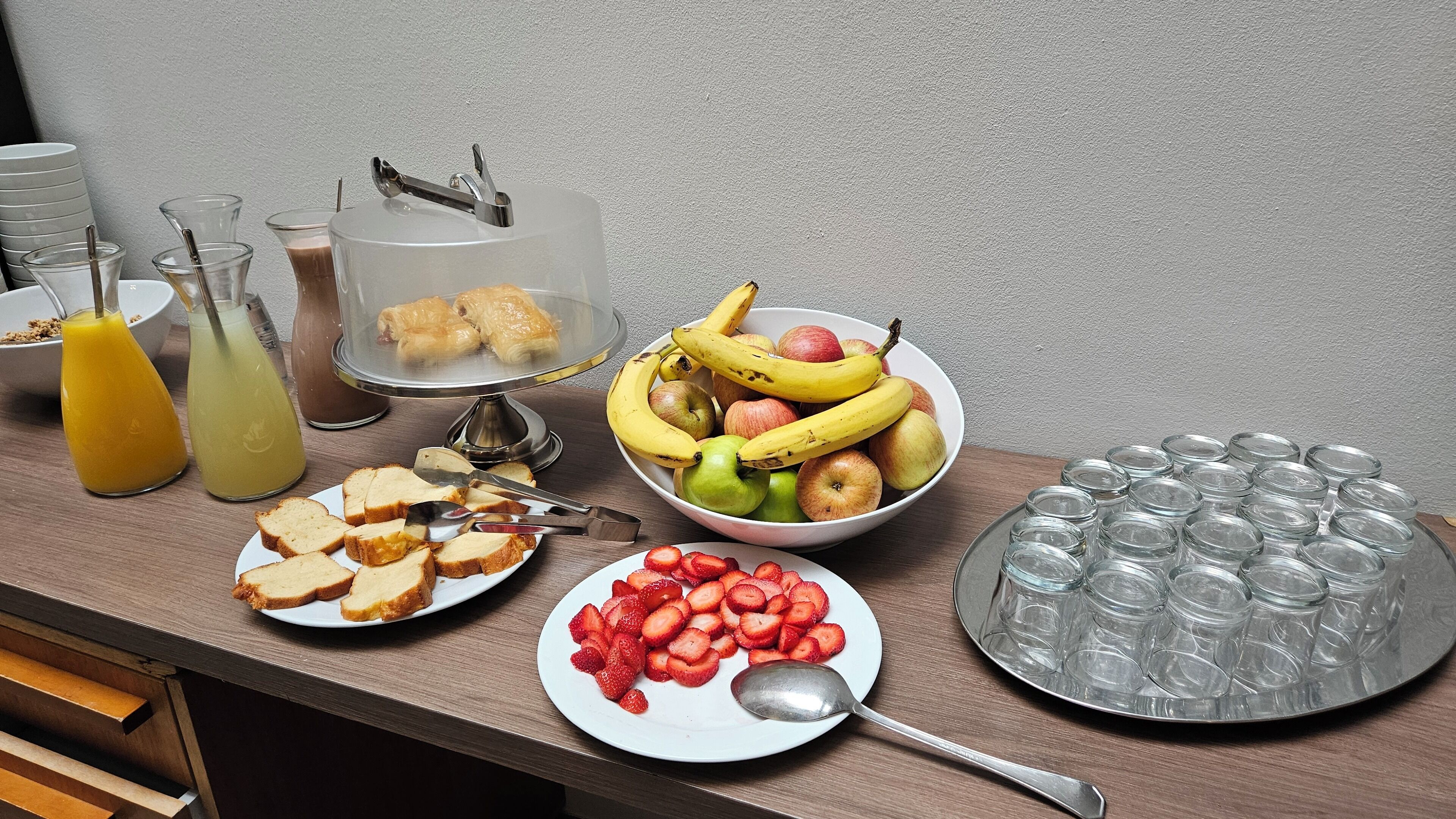 Un bufé de desayuno con pan en rodajas, fresas, un tazón de fruta y vasos de jugo.