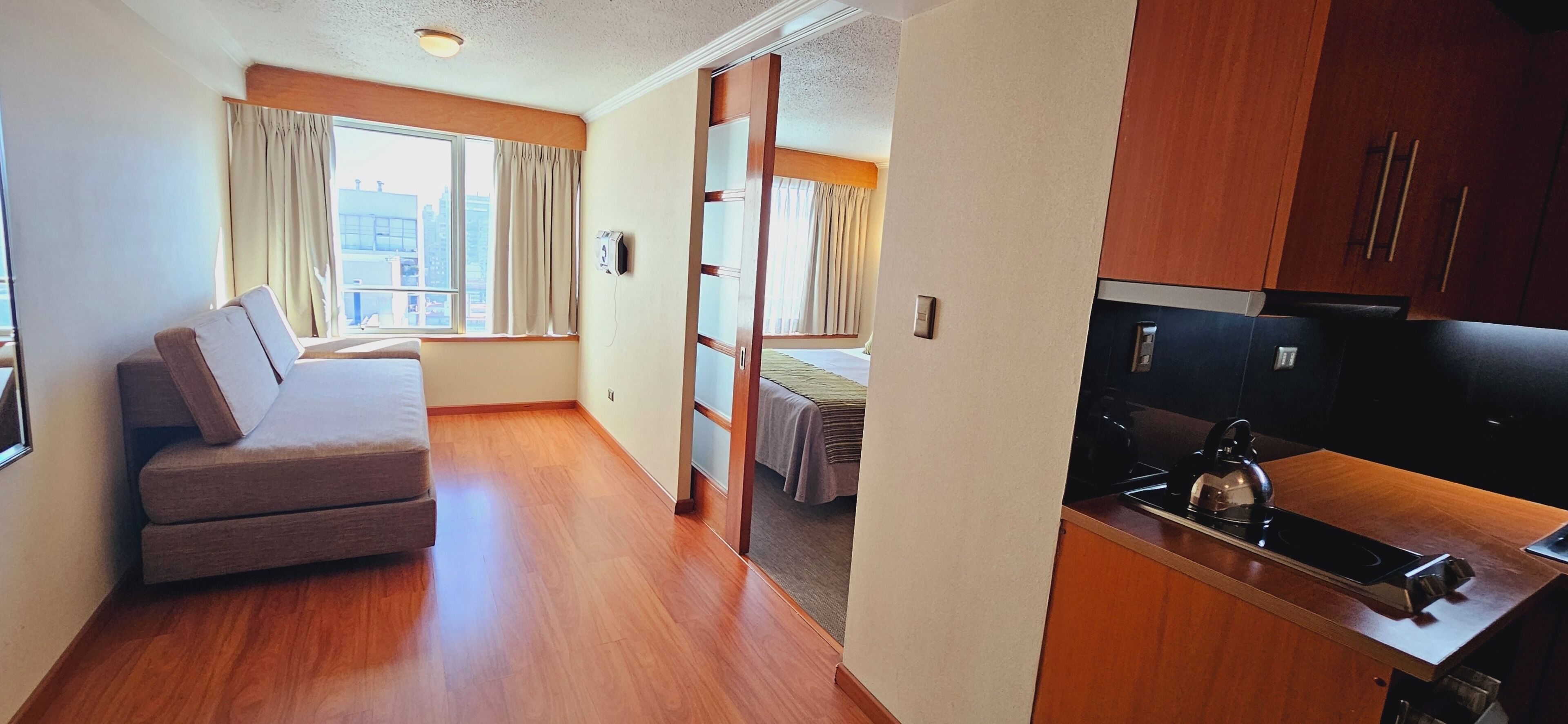 A compact hotel room with a sofa, a bed, a kitchenette, and a view of the city.