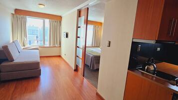 A compact hotel room with a sofa, a bed, a kitchenette, and a view of the city.