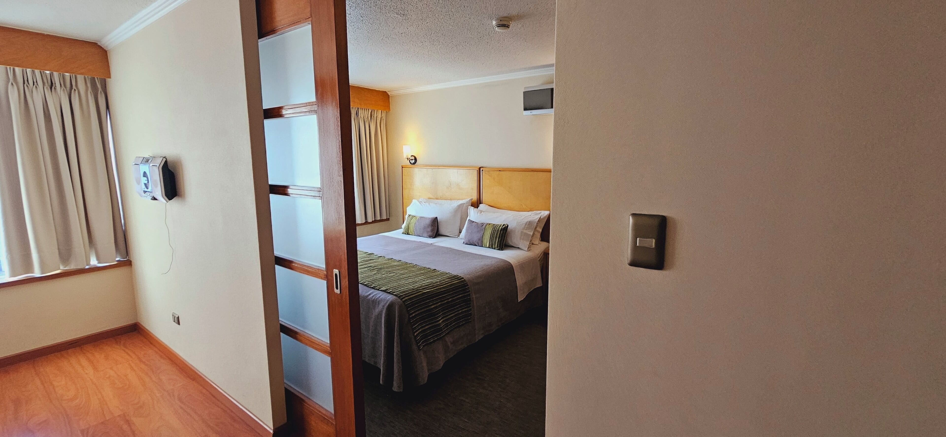 A hotel room with a bed, a bedside table, a wall-mounted TV, and a window with curtains.
