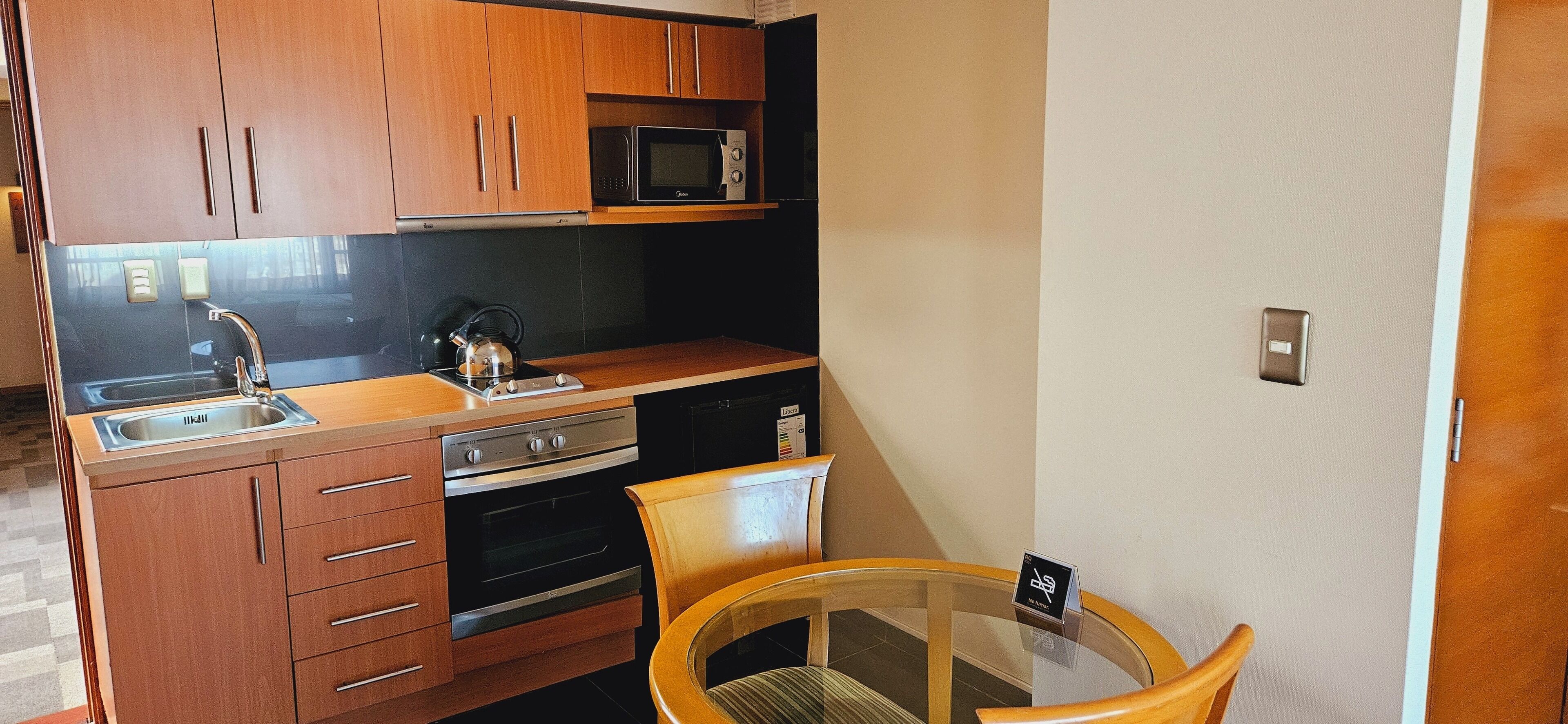 A compact kitchen with wooden cabinets, a microwave, a kettle, a sink, and a small dining table with a chair.
