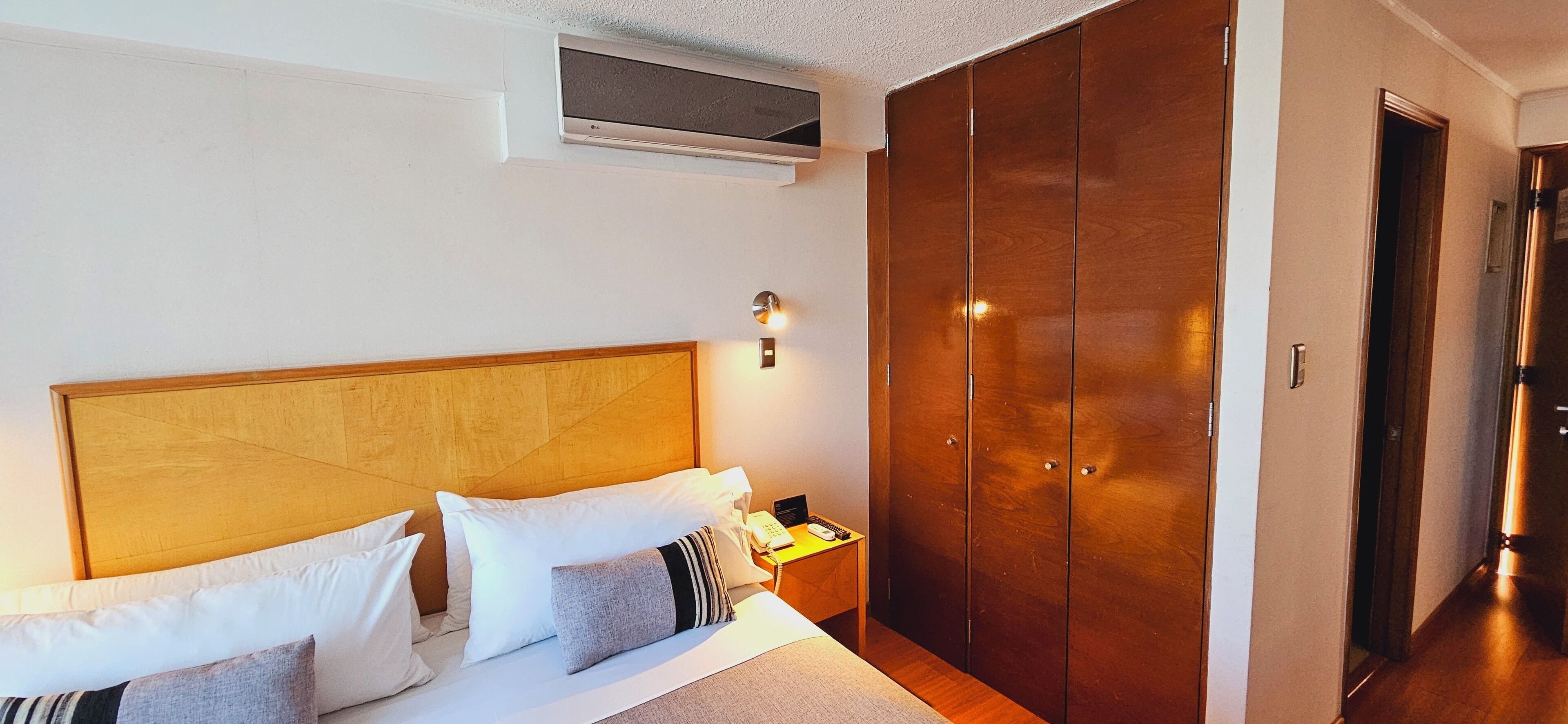 Una camera d'albergo con una testiera in legno, un letto con lenzuola bianche e numerosi cuscini, un comodino con lampada e telefono, e un ampio armadio.