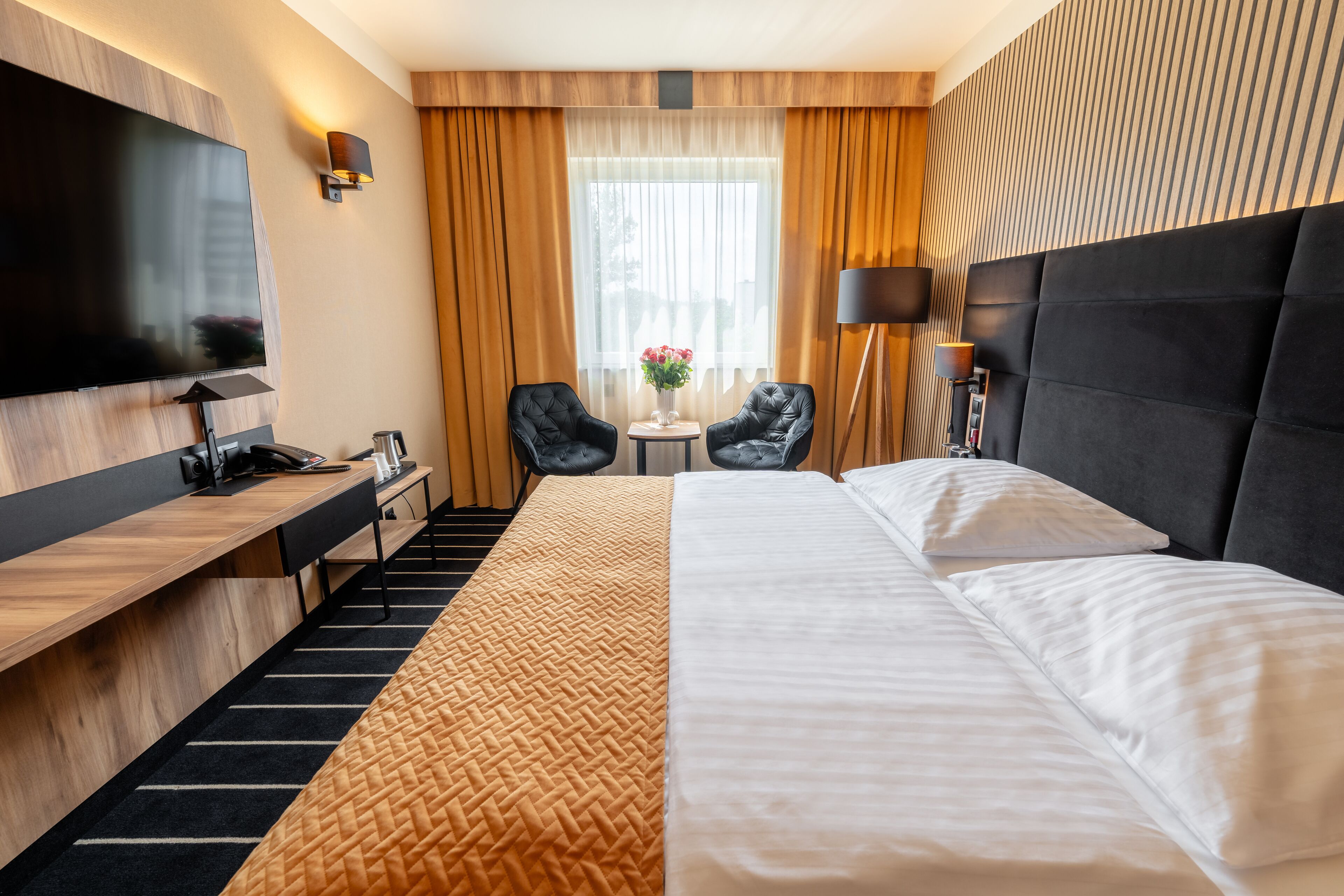 A hotel room with a large bed, a desk with a flat-screen TV, a small table with chairs, and a large window with curtains.