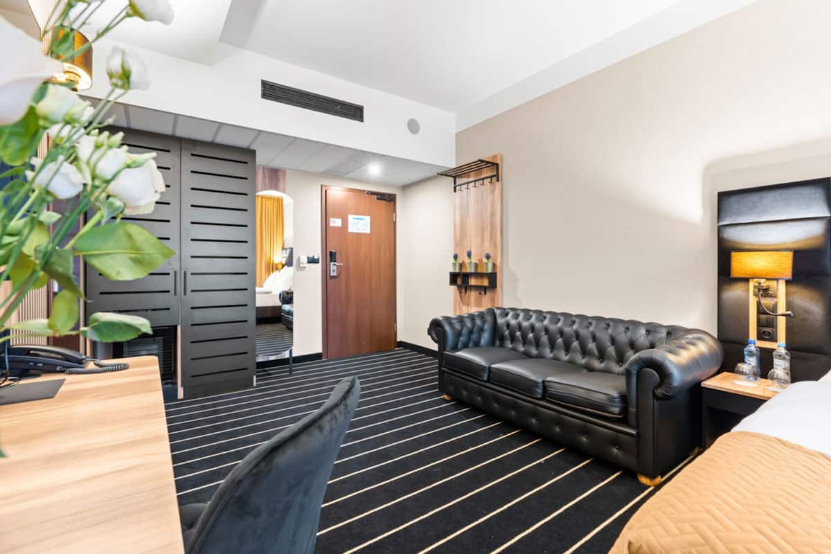 Een hotelkamer met een zwart-wit gestreept tapijt, een zwarte leren bank, een houten bureau met telefoon en een bed met een beige deken.