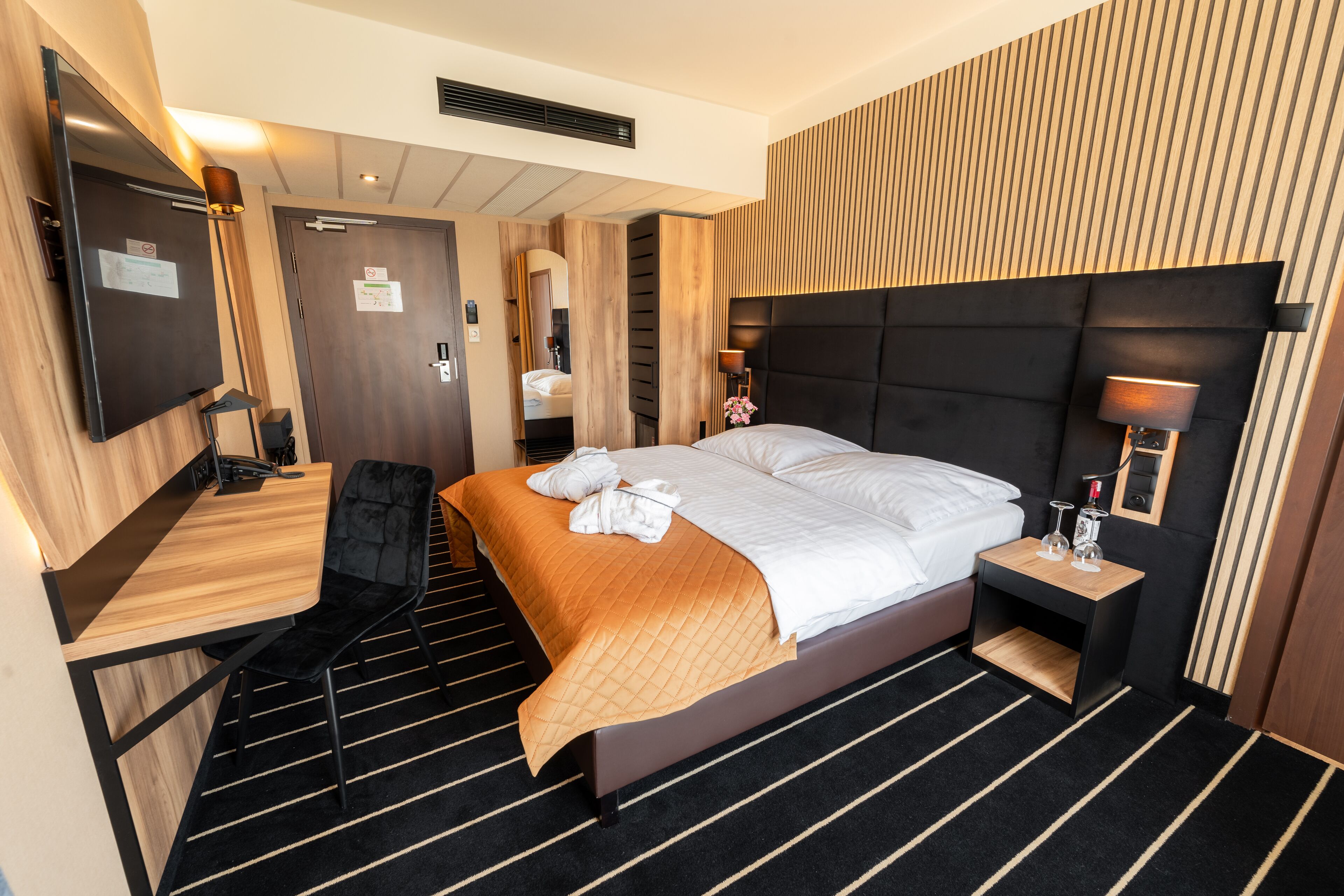 A modern hotel room with a large bed, a desk, a chair, a TV, and a bedside table with a lamp.