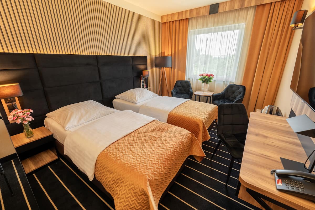 Een hotelkamer met twee bedden, een bureau, een stoel, een lamp, een kleine tafel met bloemen en een groot raam met gordijnen.