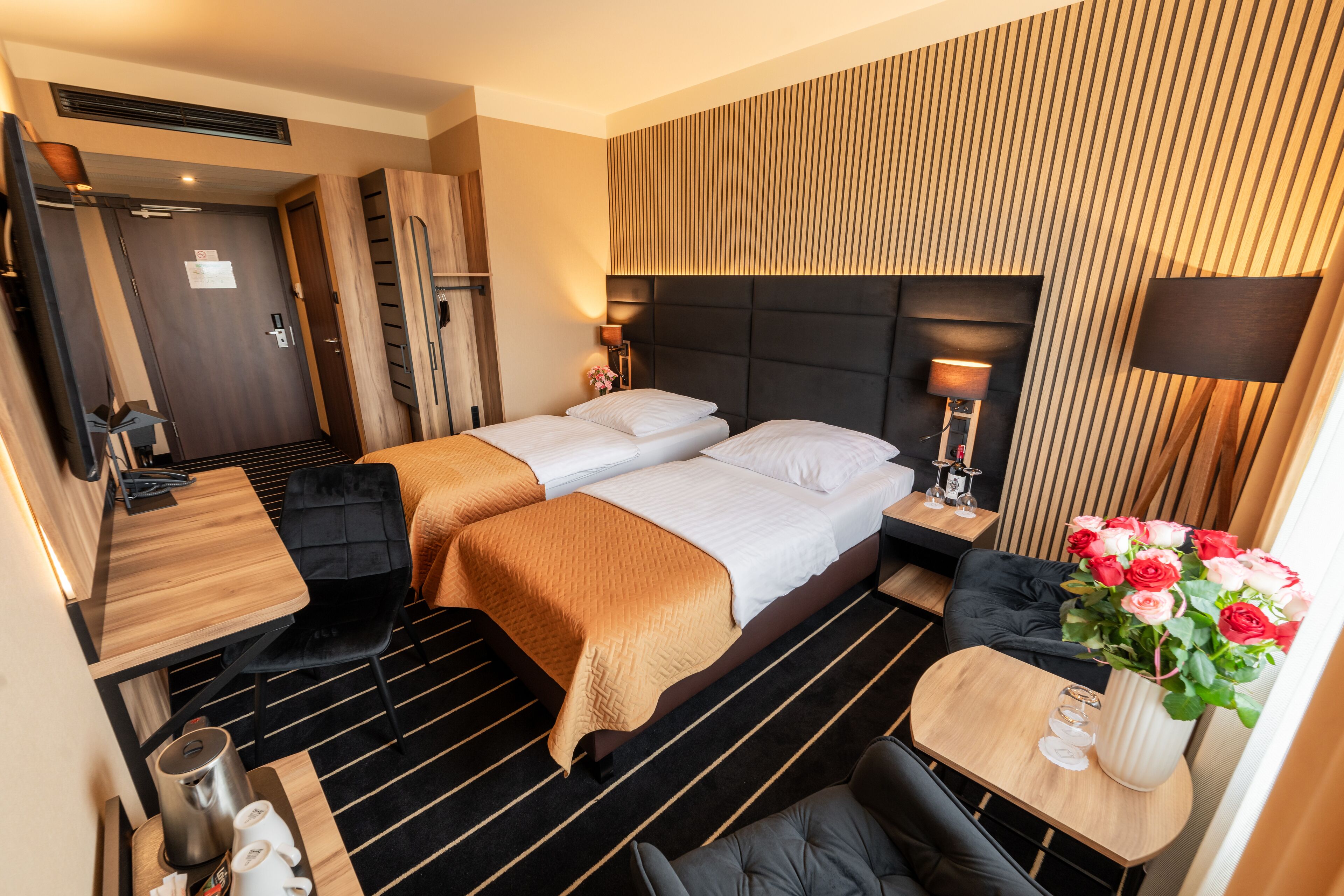 A hotel room with a bed, bedside tables, a desk, a chair, a coffee maker, and a vase with flowers.